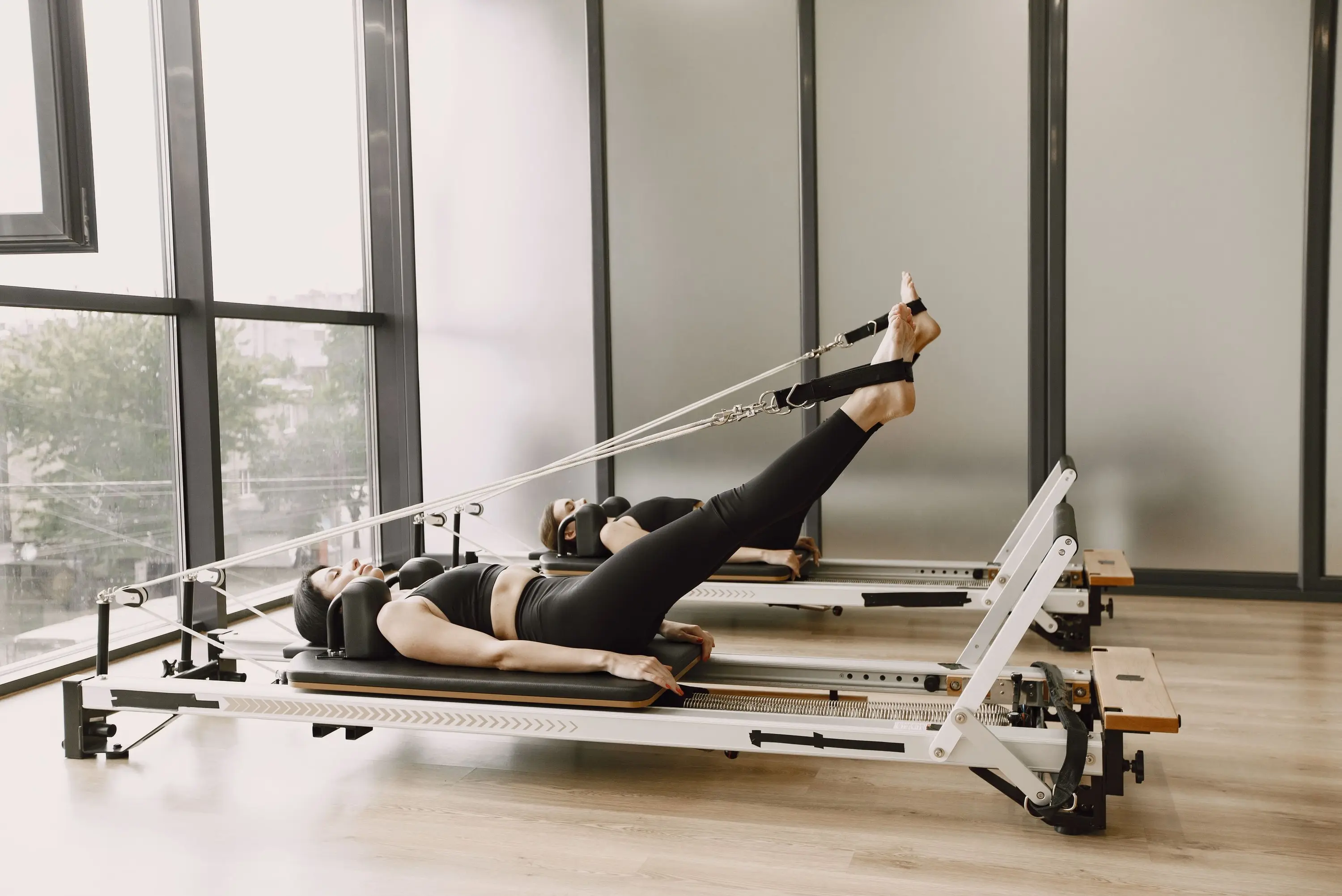 Two people lying on reformers working through footwork in a bright studio, feet in the straps, springs loaded light.