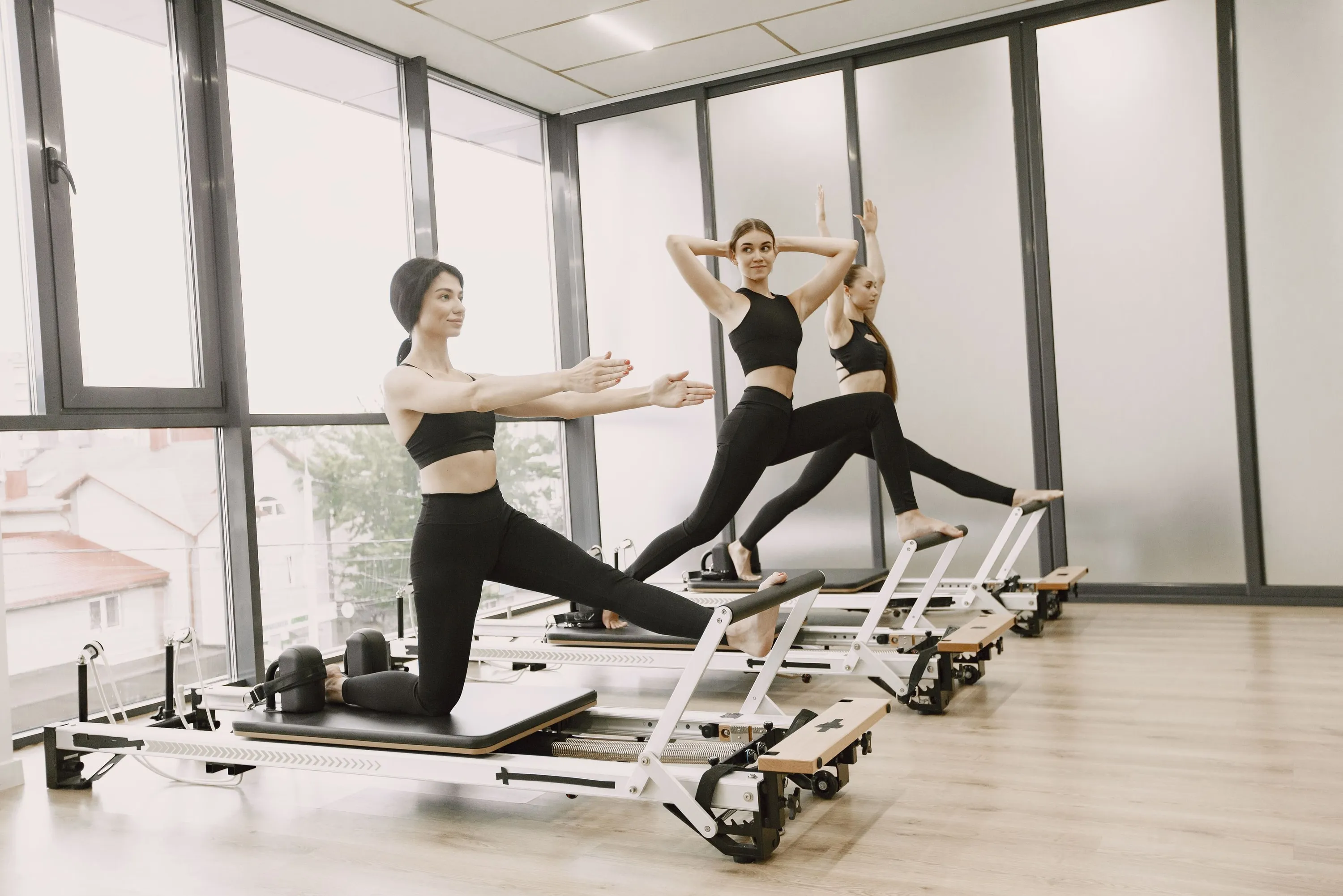 Three people mid-sequence on reformers in a bright studio, one lunging on the carriage with arms raised.