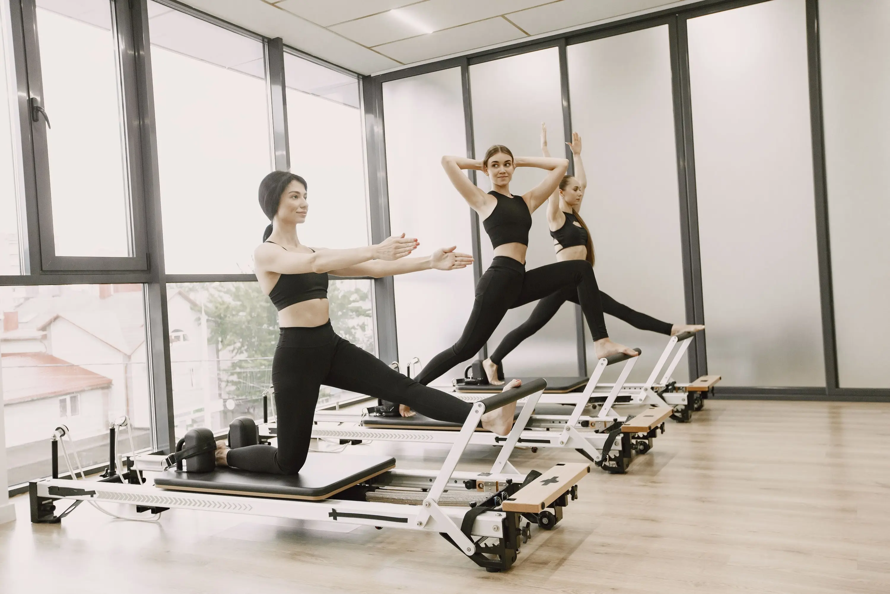Three people mid-sequence on reformers in a bright studio, one lunging on the carriage with arms raised.