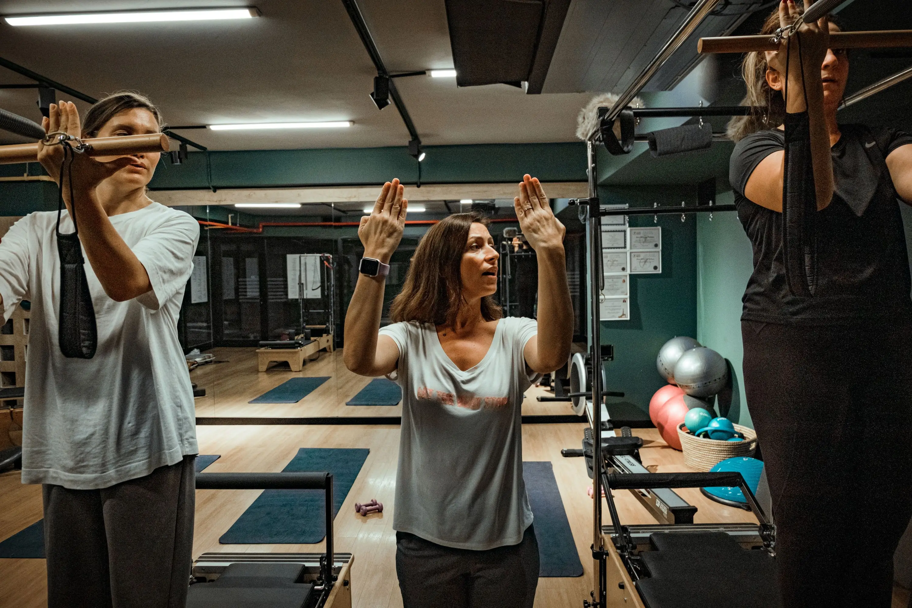 Priya cueing a class with both arms raised, two clients working at the tower bars either side of her.