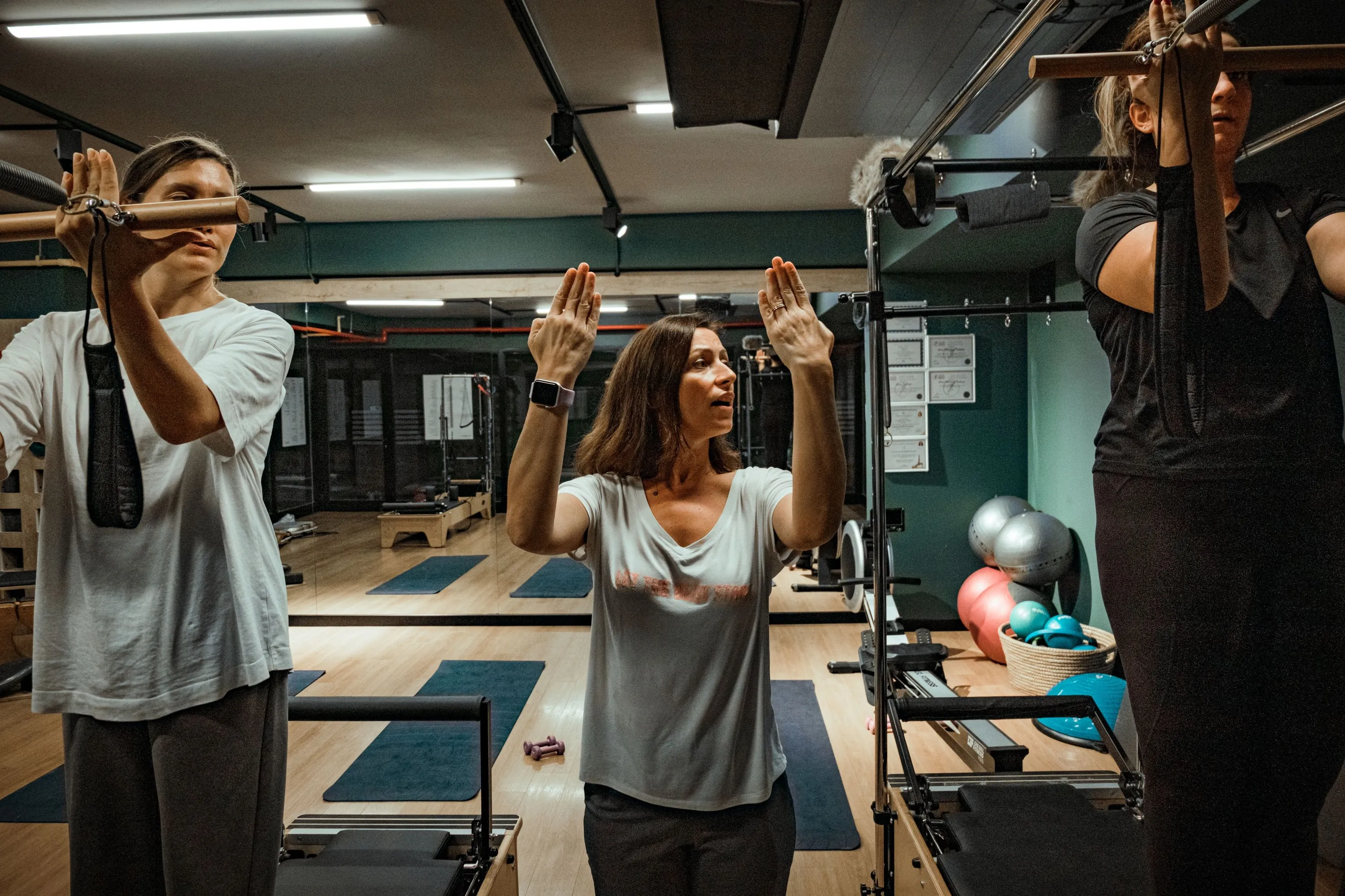An instructor cueing a movement with both arms raised while two clients work at the tower bars either side of her.
