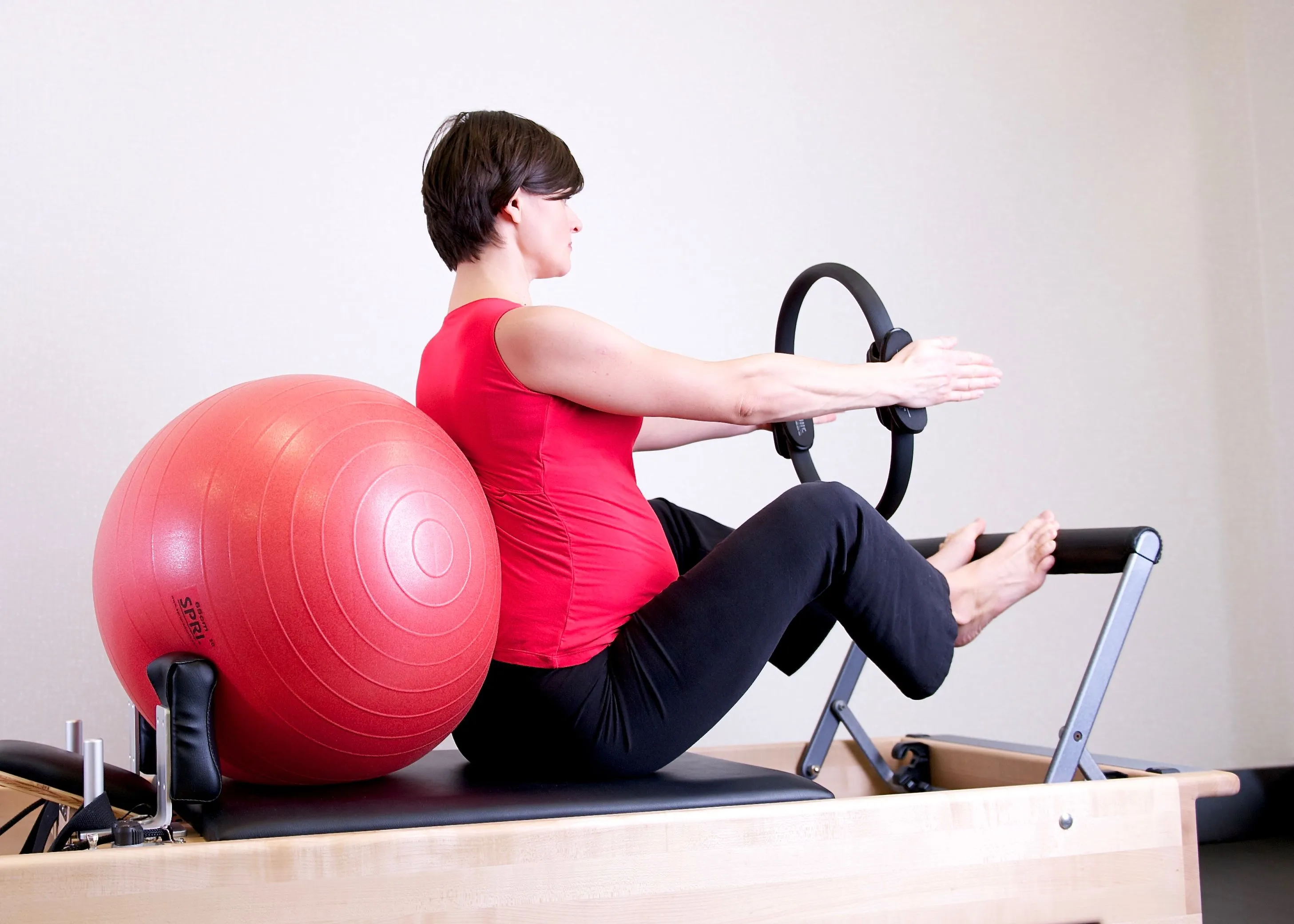 A pregnant woman sitting on a reformer carriage supported by a large ball, working with a magic circle held at chest height.